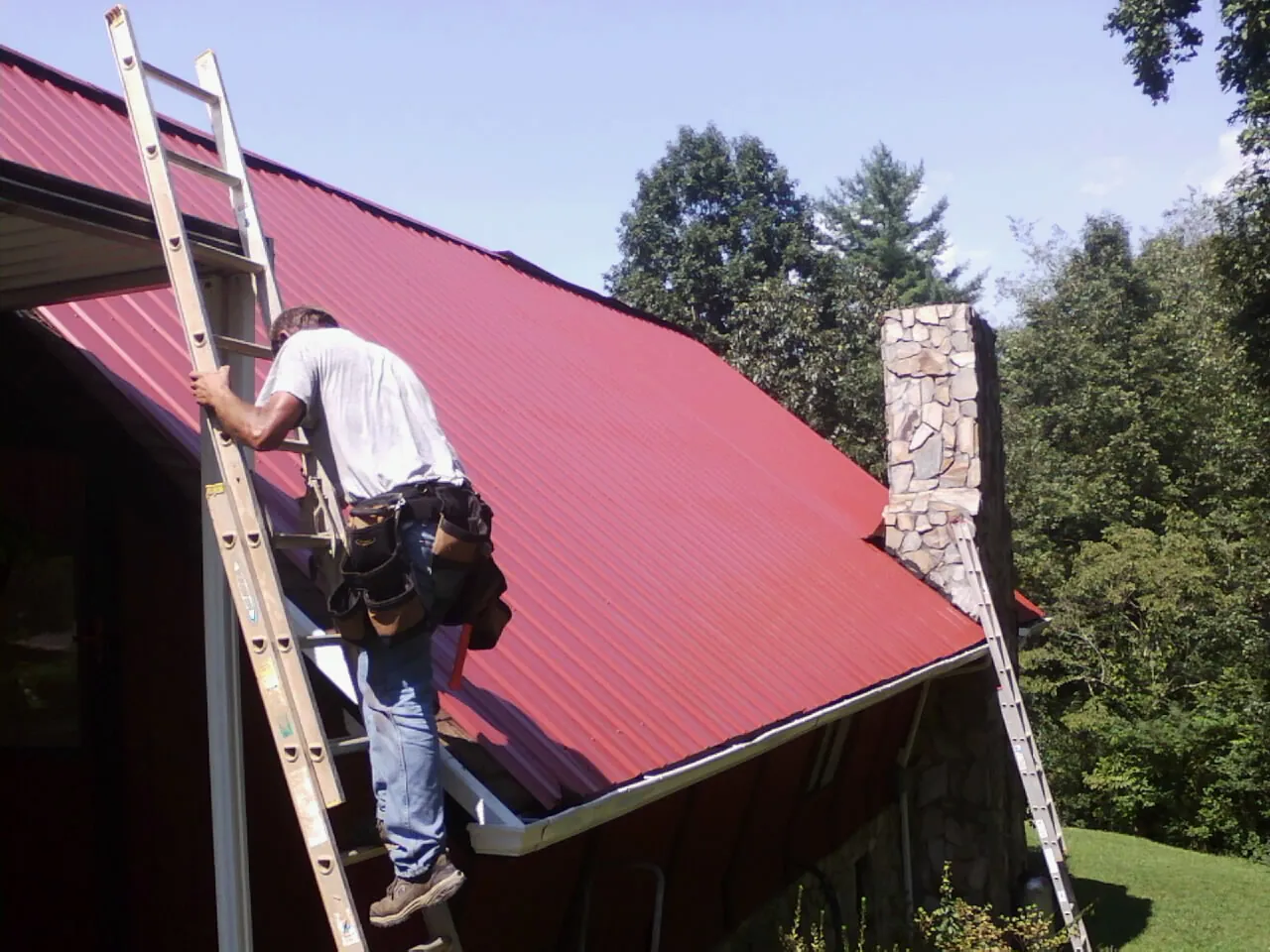 Roofer on a ladder beside a red metal roof and stone chimney — Soffit & Fascia Repair by Roofing Brothers in Caliente